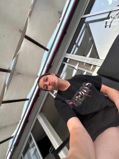 The image shows a young person from the chest up, taking a selfie. They are wearing a black t-shirt with a red and white graphic design on the front. Their face is angled towards the camera, and they are making a pouty expression. The background appears to be outdoors, with a view of a building with large windows and some sort of awning or canopy structure. The lighting suggests it is daytime, possibly sunny. The image is taken from a low angle, and the person's arm is extended to capture the selfie. The person's skin tone is fair, and they have dark hair. There is some visible text on the t-shirt, but it is partially obscured and not entirely legible. The setting appears to be a balcony or terrace overlooking a built environment.