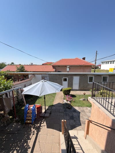 The image depicts a sunny, outdoor scene in the city of Dolni Dabnik, Bulgaria. The viewpoint is from an elevated position, looking down into a backyard area. A light-colored umbrella is open in the center of the frame, casting a shadow. To the left of the umbrella, a blue and yellow plastic children's slide is visible. A small bed of pink flowers is situated on the grass to the right of the slide. Beyond the backyard, a single-story building with a red-tiled roof is prominent, with concrete walls and white doors and windows. Further in the background, a larger white building with a multi-paned facade can be seen, along with utility poles and overhead wires. The ground is paved with square tiles in some areas and covered with grass in others. A black metal railing runs along the edge of the elevated vantage point. The sky is clear and bright blue, indicating fair weather.