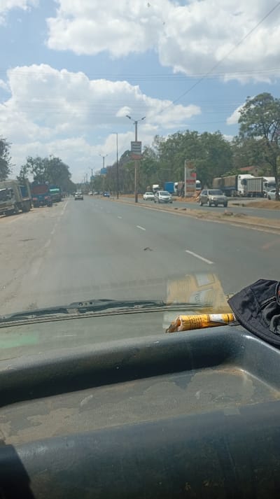A driver's perspective of a wide, mostly empty highway under a cloudy sky. Various vehicles, including trucks and cars, are sparsely scattered along the road and parked on the shoulder. A roadside sign and trees are visible in the distance. The vehicle's dusty dashboard is in the foreground, with a discarded wrapper and a black cap resting on it.