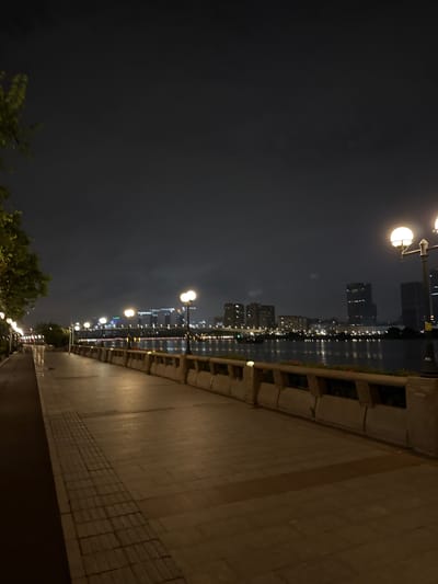 A nighttime view of a waterfront promenade in Tianhe District, China, reveals a paved walkway bordered by a low wall with planters. Streetlights with glowing globes illuminate the path, casting warm light on the textured paving stones and a darker, red-hued path to the left. Across the water, a city skyline is visible, featuring illuminated buildings and bridges. A few trees are present on the left side of the promenade, their leaves dark against the night sky. The water itself reflects the distant city lights. The scene is tranquil and deserted.