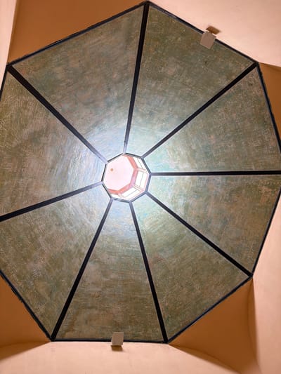 The image shows an interior view looking upwards at a domed ceiling with an octagonal opening at the apex, allowing natural light to filter in. The domed ceiling is divided into eight triangular sections, each painted with a mottled green and cream texture, bordered by dark lines. Two small white square lights are mounted on the ceiling. The surrounding walls are a warm peach color. This architectural feature is located in the Templo Masónico building in San Cristóbal de La Laguna, Spain.