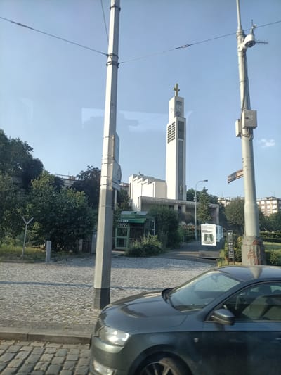 This is a medium-angle, slightly tilted, outdoor, daytime, eye-level shot of the Church of St. Wenceslas in Prague, Czechia. The church, a prominent white structure with a tall, slender tower topped by a gold cross, is the central subject. The tower features a clock face and vertical decorative elements. The church building itself is rectangular with a smaller rectangular spire extending from its side. To the left of the church, a modern, light-colored building with numerous windows is visible.
In the foreground, the hood and front passenger side of a dark gray car are visible, obscuring the lower part of the scene. The car is parked on a cobblestone street. To the left of the car, there is a sidewalk also paved with cobblestones, and beyond that, a patch of grass and trees.
Two tall, gray, industrial-looking poles with overhead wires are prominent in the foreground and middle ground, suggesting the presence of public transportation, likely a tram. The pole on the right has a street sign attached to it, reading "STRESOCKEHO NA MYTI". A smaller, green kiosk-like structure with glass doors is located near the base of the church complex, and a white sign with a drawing of a woman in a dress is displayed on an easel to the right of the kiosk.
The sky is a clear, light blue. The overall impression is a sunny day in a city environment, with a mix of modern architecture and historic paving.