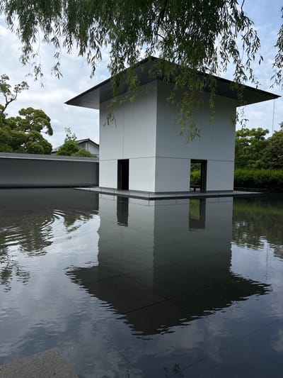 A minimalist, modern, white, cube-like building with a wide, dark grey overhanging roof stands centrally within a dark, still reflective pool of water under an overcast sky. The building features two dark, rectangular open doorways on the side facing the viewer, which are clearly reflected in the water below. The water's surface exhibits subtle ripples, particularly on the left side, slightly distorting the reflections of the sky and surrounding foliage. The building itself is situated on a dark, tiled platform that extends into the water.
In the upper portion of the frame, weeping willow branches with slender green leaves hang down, partially obscuring the building's roof and the sky. To the left of the building, a long, low, grey wall runs horizontally, behind which lush green trees with dense canopies are visible. Further in the background, a traditional Japanese-style dark tiled roof is discernable amongst the trees. To the right of the building, dense green bushes and trees form a continuous backdrop. The reflections in the water accurately mirror the building's white walls, dark doorways, and the overhead sky and willow branches. No people are visible in the scene. This architectural scene is characteristic of the D.T. Suzuki Museum in Kanazawa, Japan.