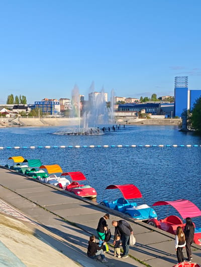 The image depicts a waterfront scene in Vinnytsia, Ukraine, featuring a body of water with a fountain in the center. The fountain is spraying water upwards, creating a rainbow effect. A line of colorful paddle boats is docked along a concrete embankment. A group of people, including adults and children, are gathered near the paddle boats. The background includes buildings and trees. The sky is clear and blue. The overall scene suggests a leisure or recreational area within the city.