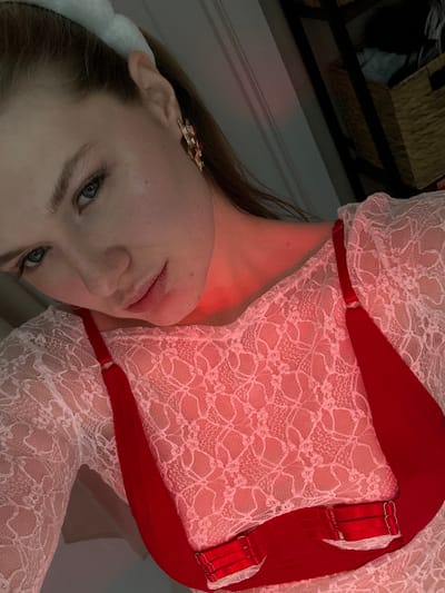 A woman in a white lace bodysuit with red straps poses. She wears an ornate gold earring and a white headband. The image is dimly lit, with a red cast on her face and neck. Part of a woven basket is visible in the background. The photo is taken at an angle, making the woman appear sideways.
