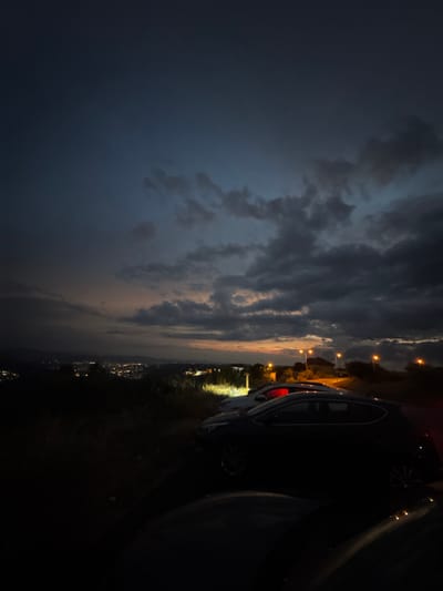 This vertical, low-light photograph captures a scene at dusk or dawn, overlooking a distant, illuminated town that stretches across the horizon. The sky is dominated by dark, dramatic clouds with streaks of orange and yellow where the sun is setting or rising. In the foreground, several cars are parked on what appears to be a dirt or gravel surface, possibly a viewpoint or parking area on a hillside. The closest car, a dark hatchback, is the most prominent, its silhouette sharply defined against the faint light. Other vehicles are visible behind it, one with its brake lights illuminated a bright red. Streetlights dot the landscape, some with a warm orange glow, illuminating the immediate surroundings and hinting at the urban expanse below. The environment is somewhat wild, with dark vegetation and dry grass visible on the left side of the frame. The overall impression is one of quiet observation, with the illuminated town providing a stark contrast to the darkening natural landscape and the parked vehicles. The location of Arbanassi, Bulgaria, is mentioned in the prompt, suggesting the view is from a vantage point above the town, offering a panoramic perspective.