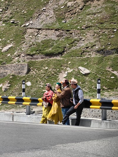 Four individuals are standing on the side of a road in Burwa, India, with a steep, grassy mountainside behind them. The individuals are a mix of genders and are dressed in traditional clothing, including hats with distinctive patterns. They are gathered near a yellow and black guardrail that separates the road from the embankment. One person is holding a phone, possibly taking a photo or video. The environment appears to be a rural or mountainous area, with visible rocks and sparse vegetation on the slope. The road surface is asphalt, and there is a concrete barrier along the edge of the road.