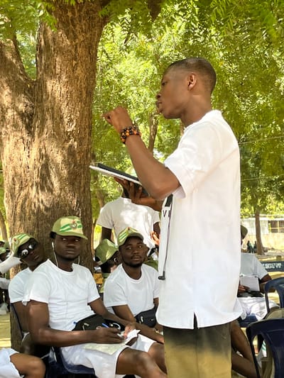 An outdoor instructional session is depicted in Maiduguri, Nigeria. A young Black male, wearing a white short-sleeved shirt, dark trousers, and beaded bracelets on his right wrist, stands gesturing with his raised right hand while holding a black notebook or tablet in his left. He faces slightly left, addressing an unseen audience. Several young Black males are seated on blue plastic chairs to his left, forming an audience. They are uniformly dressed in white short-sleeved t-shirts and olive green camouflage caps featuring a distinct emblem on the front. One individual in the foreground is actively taking notes on a white sheet of paper with a black pen, while another beside him wears sunglasses on his cap. A large tree trunk is prominent in the mid-ground, with dense green foliage filling the background, indicating a sunny, open-air environment. Faint building structures are visible in the far background.