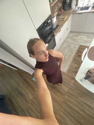 A woman in a maroon dress is taking a selfie in a well-lit room with wooden floors. She is standing and looking into the camera, with her right arm extended. The background shows a modern kitchen with white cabinets and a black countertop. On the countertop, there are various kitchen appliances and utensils. A dining table with a remote control is visible in the upper right corner. The room appears to be a residential interior in Подшивалово, Russia, and the time of day is likely daytime due to the bright lighting. The woman's expression is neutral. No visible text is present in the image.