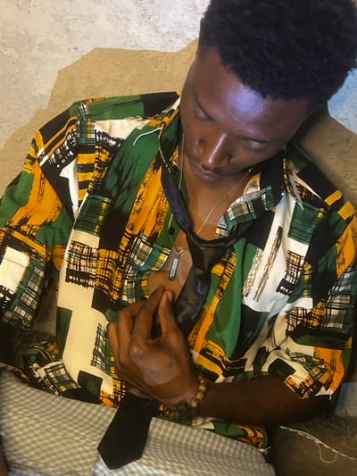 A young Black male is depicted from an elevated perspective, seated or leaning against a surface. His head is tilted downwards and slightly to his right, with eyes cast down or closed. He wears a short-sleeved collared shirt featuring an abstract pattern of green, yellow, black, and white brushstrokes. An untied black necktie is draped around his neck, resting against his chest. A silver-colored chain with a rectangular pendant hangs around his neck. His right hand is positioned on his chest, lightly gripping the black tie. A bracelet made of wooden beads is visible on his right wrist. He is wearing light-colored, possibly checkered, lower garments. The immediate background behind his head and left shoulder is a light tan, textured surface, while a darker, indistinct textured surface is visible to his right. The scene occurs in Bar Gada, Nigeria.