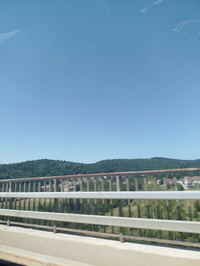 This image depicts a view from a bridge in Villards-d'Héria, France, overlooking a densely forested hillside under a clear blue sky. The foreground is dominated by the bridge structure, with horizontal metal railings and a concrete walkway. Through the railings, a verdant landscape unfolds, characterized by a thick canopy of green trees covering rolling hills. In the distance, nestled amongst the trees, are the rooftops of houses, suggesting a small village or settlement. The overall scene conveys a sense of rural tranquility and a bright, sunny day. There are no people visible in the image.