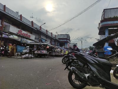 A black motorcycle is parked on the street in front of a building, partially obscured by the motorcycle's large, silver, front tire. The front end of the motorcycle is pointed towards the right side of the image. The street is made of asphalt. The camera is positioned on the ground, looking upwards, so the buildings in the background appear upside-down. The buildings have red and white trim. There are many people walking in the distance, obscured by the motorcycle and the perspective of the image. The sky is cloudy and the sun is shining in the top right corner. The building in the background has a red sign that says "Raja" on it. The scene is located in Kecamatan Lawang, Indonesia, at latitude -7.83, longitude 112.697.