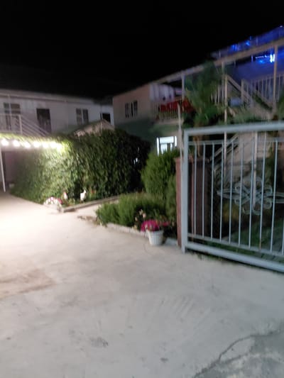 The image depicts an outdoor scene at night, likely in Ақши, Kazakhstan. The foreground features a paved walkway that extends into the distance. To the left of the walkway, a white building with multiple windows is visible, its facade illuminated by soft lights. There are balconies with white railings on the building. Adjacent to the building, a lush green hedge or dense foliage forms a natural barrier. In front of the hedge, a white flower pot contains vibrant pink flowers, adding a splash of color to the scene. A metal fence with horizontal bars, possibly part of a balcony or railing system, is prominent in the upper left section of the image. The overall ambiance suggests a peaceful evening setting. The time of day is clearly night, indicated by the darkness and artificial lighting. The weather appears calm. No people are visible in the image, and there is no visible text.