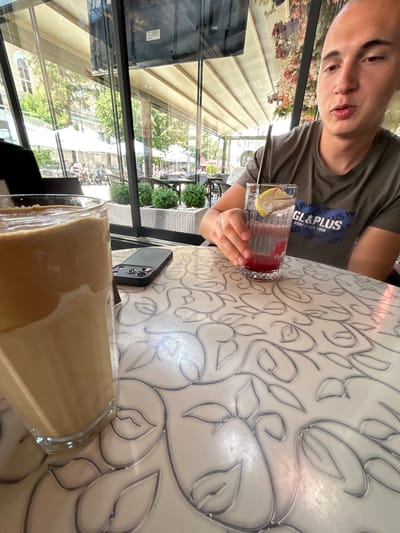 The image depicts a young man sitting at a table in an outdoor cafe, possibly in Vratsa, Bulgaria, as suggested by the prompt, though this cannot be definitively confirmed from the visual information alone. He is holding a glass with a red drink and a lemon slice. In the foreground, a tall glass filled with a layered coffee drink, likely Dalgona coffee, is placed on the table. A smartphone case is also visible on the table next to the coffee. The background shows a street scene with trees, buildings, awnings, and people. The cafe appears to be located along a street with a pedestrian walkway.