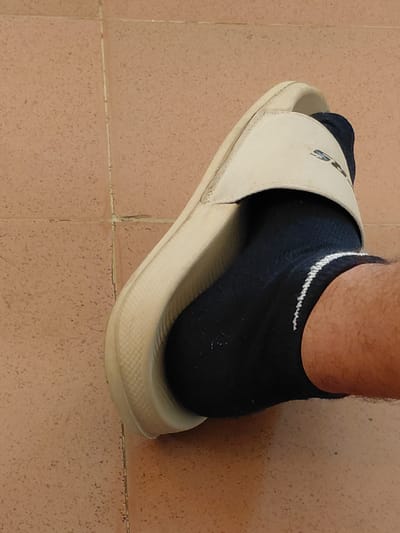 A person's left foot, wearing a black sock with a white stripe and a light-colored slide sandal, is positioned on a tiled floor. The floor is made of square, reddish-brown tiles with visible grout lines. The sandal is open-toed and has a logo on the strap. The image is taken from a close-up, slightly elevated perspective. The setting is indoors, and the lighting is neutral, casting soft shadows. The specific location in Santa Ana, Venezuela, cannot be determined from the visual information provided.