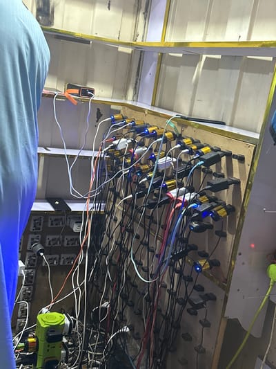 In Durumi, Nigeria, an individual wearing a blue garment stands to the left of the frame, partially obscuring a wall covered with numerous electrical outlets and chargers. A power bank sits on a shelf above the outlets. The wall itself is a makeshift structure, with exposed wooden panels and a corrugated metal or plastic backing. Numerous charging cables, predominantly black and white with some red and blue accents, are plugged into a dense array of multi-port USB chargers. Many of these chargers have small blue indicator lights illuminated, suggesting active charging. A bright green power strip with multiple sockets is visible at the bottom left, also connected to various devices. The overall environment appears to be an indoor space, possibly a workshop or a charging station, characterized by a high concentration of electrical equipment and wiring.