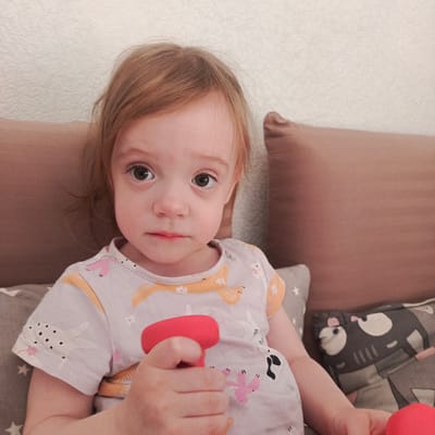 A young child with reddish-blonde hair is holding a red object, possibly a toy, in their right hand. The child is sitting against a backdrop of brown and gray pillows. The child is wearing a light-colored t-shirt with animal designs. The child has a solemn expression. The location is Марковский, Russia.