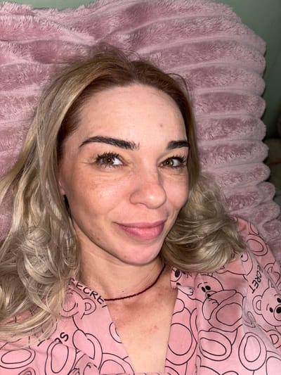 This is a close-up, overhead shot of a woman lying on a pink, fuzzy blanket. She has blonde hair with lighter streaks, and her head is tilted to the side. She is wearing a pink pajama top with a repeating pattern of cartoon teddy bears and the word "SECRET" visible on the collar area. A thin, dark red beaded necklace is visible around her neck. The lighting appears to be indoor, with a slight green cast to the background, possibly indicating a wall or other room element. The woman's eyes are open and looking towards the camera, with a soft, somewhat contemplative expression. Her skin has a warm tone, and there are visible freckles and some redness on her cheeks and neck. The image is a portrait that focuses on the woman's face and upper body, emphasizing her relaxed state on the soft blanket.