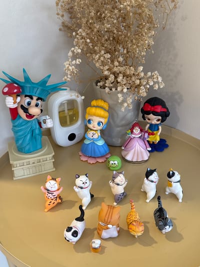 This media file is a top-down, rotated shot of various figurines placed on a light brown, circular surface, possibly a tray or table. The setting appears to be indoors, with a neutral, off-white wall visible in the background.
The main subjects are a collection of small figurines. These include:
* A figure of Super Mario dressed as the Statue of Liberty, holding a mushroom.
* A doll resembling Cinderella, holding a clock.
* A doll resembling Snow White, holding another doll with red hair.
* Several cat figurines, some lying down, others in playful poses, with various patterns including orange tabby and black and white.
* A small green frog figurine.
* A white humidifier or diffuser device.
* A small arrangement of dried, light-colored flowers.
The figurines are arranged haphazardly, with no clear narrative or activity taking place. The overall impression is of a collection of decorative items. There is no visible text in the image. The lighting suggests it might be daytime, possibly with ambient light from a window. The location context of Suthep, Thailand, does not provide specific visual cues within the image itself.