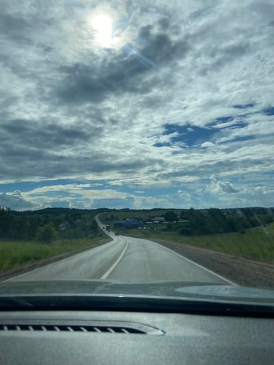 The media file shows a scenic view of a road curving through a rural landscape in Забегалово, Russia. The sun is shining brightly through a dramatic sky filled with a mix of dark and light clouds, suggesting a partly cloudy or overcast day. The road is paved with white dividing lines and is bordered by lush green fields and trees. In the distance, a small cluster of buildings, likely a village, can be seen nestled amongst the trees. The image appears to be taken from inside a vehicle, as indicated by the presence of a car interior on the right side of the frame, including a dark-colored dashboard and a vent. The overall mood is one of peaceful travel through a picturesque countryside. There is no visible text in the image.
