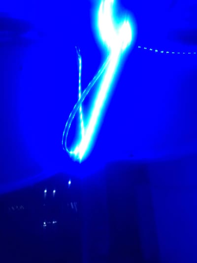 The image depicts a dark, blurry scene dominated by bright blue and white light streaks, suggesting long exposure photography or movement of light sources. The background is a deep, saturated blue, with some darker areas at the bottom suggesting a surface or floor. In the upper right portion of the image, a series of small, evenly spaced blue lights are visible, forming a line that trails off into the distance. A prominent, thick streak of bright white and blue light curves across the center of the frame from the top left to the bottom right, with thinner, more intricate light trails wrapping around it, possibly indicating movement of a light-emitting object or person. The overall impression is abstract and dynamic, with no discernible landmarks or specific environmental details to place it in Howayi, Nigeria, or any other specific location. The image is highly processed, with the lights appearing to be the sole focus of the composition, obscuring any contextual information.