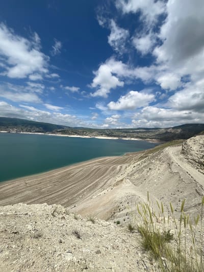 A vertical shot of the Chirkey Reservoir in Chirkey, Russia shows a large body of water bordered by rocky terrain on the right and rolling hills with clouds in the background to the left. The rocky terrain has sparse vegetation, while the hills have more, darker vegetation. The sky is blue with scattered cumulus clouds. The water appears to be a slightly greenish-blue color.