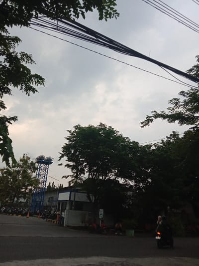 The image displays an outdoor scene in an area of Semarang, Indonesia, under an overcast sky. The foreground features a dark asphalt road. In the left midground, a tall blue lattice water tower supports two circular tanks. Adjacent to the water tower, a low-rise building with a white facade and blue trim is visible. In front of this building, a paved lot contains numerous parked motorcycles and several orange traffic cones. The center and right midground are dominated by dense, dark green trees. Beneath these trees on the right, two individuals are seated on a low structure or bench. A red scooter is parked nearby. On the asphalt road to the far right, a black scooter with two occupants, the operator wearing a light head covering, travels from right to left. Overhead, multiple dark cables span horizontally across the sky, partially obscured by dark tree branches originating from the upper left.