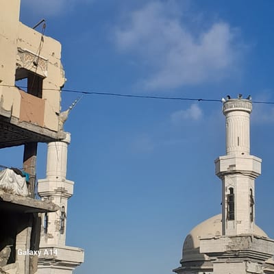 The image shows two minarets against a bright blue sky with scattered clouds. The minarets appear to be part of a mosque, one of which is intact and the other is in ruins. The intact minaret is tall and cylindrical with decorative elements, topped with several lights. The ruined minaret is fragmented, with exposed rebar and broken concrete.
Below the minarets, the remnants of a building are visible, also in a state of disrepair. The scene suggests a context of destruction or damage. A thin black wire runs diagonally across the image, bisecting the sky and parts of the structures. The lighting indicates it is daytime, likely late morning or early afternoon, given the bright sunlight and clear sky. There are no people visible in the image.
The visible text in the image reads "Galaxy A14", which is a model name for a Samsung smartphone, indicating it is likely a watermark from the device used to capture the photograph. The location context provided is Khan Yunis, Palestinian Territories, which aligns with the imagery of damaged structures, possibly a result of conflict. The overall mood conveyed by the image is one of devastation and loss, juxtaposed with the enduring presence of religious architecture and the clear sky.