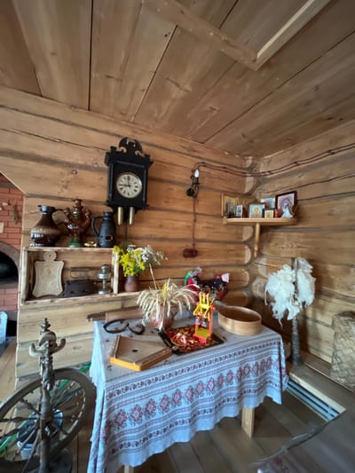 This media file captures a rustic interior scene, likely a traditional Russian dwelling in Vybolovo, Russia, given the context. The walls and ceiling are constructed from wooden planks, creating a warm, textured environment. Various objects are displayed, suggesting a collection of folk art or historical artifacts.

Prominently featured is a black, ornate grandfather clock with a visible clock face. Adjacent to it are shelves holding religious icons, a wooden bowl, and decorative items. Further to the right, a table is draped with a white cloth adorned with a red floral pattern. On this table, a kantele-like musical instrument, a wooden vase with dried wheat stalks, and other small items are arranged. A spinning wheel is visible in the lower right portion of the image, indicating a historical context related to textile production. Scattered throughout the scene are various other decorative and functional objects, including ceramic vessels and what appears to be raw wool. The overall impression is one of a well-preserved, traditional home or museum exhibit, showcasing elements of rural Russian life and culture. There is no indication of people or a specific event taking place. The lighting suggests daytime, and the interior setting is cozy and filled with historical charm.