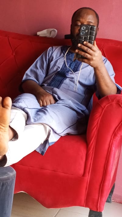 A man sits on a red sofa, facing the camera, holding a smartphone in front of his face. He wears a light blue shirt with dark blue trim and white pants. A pair of white socks rests on the sofa behind his head. His bare foot is visible in the foreground. A dark gray ottoman is positioned at the bottom left of the frame. The background wall is a reddish-pink color.