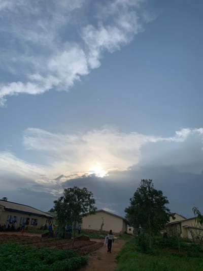 The image depicts an outdoor scene with a bright sky filled with dramatic clouds, some illuminated by the sun, which is partially obscured. The sun's rays are visible breaking through the clouds, suggesting it might be late afternoon or early morning. The scene includes a dirt path running alongside a building, with lush green vegetation on the right side of the path. Two buildings with light-colored exteriors and dark roofs are visible.
Several people are present. One person is walking on the path away from the camera, wearing dark clothing and carrying something in their hands. Another individual, wearing a blue shirt and dark pants, is standing near the first building. Further down the path, a few more people, some in blue attire, are visible near the second building. There are posters or papers displayed on the exterior wall of the second building. A tree with full foliage stands between the two buildings.
The overall atmosphere appears calm, with the natural elements of sky, clouds, and greenery dominating the composition. The location is identified as Kuru Karama, Nigeria. The weather seems to be a mix of sunny and cloudy, creating an interesting interplay of light and shadow.