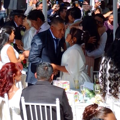 The image shows a wedding scene, likely in the city of Cerro Colorado, Peru. A bride and groom are seated at a table, surrounded by guests who are taking photos with their phones. The bride is wearing a white dress and the groom is wearing a suit. The table is decorated with flowers and glasses. The guests are dressed in various outfits. The background is blurry, but it appears to be an outdoor setting.