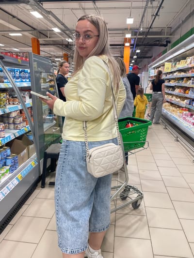 A blonde woman wearing a light yellow long-sleeved shirt, light blue denim shorts, and white sneakers stands in a supermarket aisle. She has blonde hair and wears glasses. She holds a white smartphone in her left hand. A light cream-colored quilted handbag with a chain strap is slung over her right shoulder. In front of her is a green shopping cart with a bottle and other items. Other shoppers are visible in the background, including a man in a red cap and a woman in a black top and grey pants, as well as a young child in a yellow hooded outfit. The supermarket aisle features shelves stocked with various products, including cans and containers on the left and packaged goods on the right. The floor is tiled with a light beige and grey pattern. The location is identified as a supermarket in Vologda, Russia.