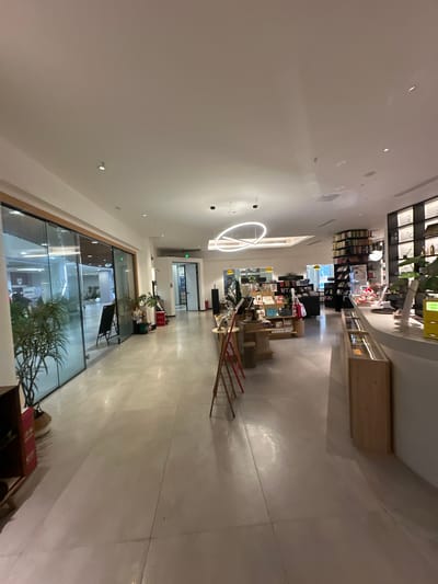 The image displays the interior of a modern, well-lit retail establishment, likely a bookstore or lifestyle store, situated in the Chongqing High-tech Zone, China. The perspective looks down a wide, open aisle towards the back of the store.

The ceiling is a clean, bright white, featuring numerous recessed spotlights and a prominent, contemporary circular light fixture that provides ample artificial illumination. The floor is a light-colored, reflective surface, possibly polished concrete or large tiles, contributing to the bright atmosphere.

On the right side of the frame, the main retail area is visible, organized with rows of dark bookshelves filled with books. Visible Chinese characters on one shelf clearly show "文學" (Wénxué), indicating a literature section. Interspersed among the shelves are various display tables and wooden stands showcasing an assortment of products, including what appear to be packaged goods, stationery, and decorative items. A distinctive long, red, multi-tiered display rack extends into the foreground on this side. Several green potted plants are strategically placed, adding natural accents to the minimalist decor.

The left side of the image features an expansive, empty walkway leading towards an area enclosed by large glass partitions. Through the glass, another interior space can be seen, which also appears modern and well-maintained, potentially another part of the store or an adjacent business.

There are no people visible in the image, suggesting a quiet time, possibly early morning or late evening, or simply a momentary absence of customers. The overall scene conveys a sense of calm, order, and modern design, characteristic of contemporary retail spaces. The lighting is entirely artificial, making it difficult to discern the time of day, but the consistent brightness creates an inviting environment.