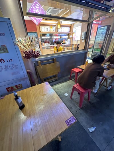 This image captures a nighttime street scene of a casual eatery, likely located in the Chongqing High-tech Zone, China. The focal point is a food stall with an open-front window revealing the interior and an adjacent outdoor dining area on the sidewalk.

Inside the establishment, visible through the window, a person in a yellow t-shirt is looking at a mobile phone while standing behind a counter. Various items associated with food preparation or serving are on the counter. Hanging from the ceiling are decorative lights and two purple diamond-shaped signs, one inverted, displaying Chinese characters, including "必吃2022" (Must Eat 2022). A hand sanitizer dispenser is mounted on the wall. To the far right, a tall, narrow glass-front refrigerator is stocked with numerous bottled and canned beverages. Above it, a red neon sign displays Chinese characters that appear to relate to "Chongqing Xiaomian" (重庆小面).

The outdoor dining area features several light-colored wooden tables and red plastic stools on a grey tiled sidewalk. In the immediate foreground, a wooden table has a glossy, possibly wet, surface with a stainless steel oil or condiment dispenser and a purple sticker with a QR code and the text "18-2". To the right of this, a person with dark hair, wearing a brown t-shirt and light-colored pants, is seated on two stacked red plastic stools, with their back mostly to the camera. They are engaged in looking at a smartphone held by another partially visible individual sitting opposite them, who is wearing light pants and white sneakers. A yellow plastic bottle rests on their table. An empty red plastic stool is next to them.

On the far left, a white panel or cabinet is visible, displaying fire safety instructions, including the number "119" (China's emergency fire number) and pictograms for fire hydrant activation and fire extinguisher use. Below this, the characters "灭火器" (fire extinguisher) are visible. Attached to this panel, a large, fan-like display of numerous candied fruit skewers (tanghulu), each wrapped in clear plastic with red sticks, is prominently featured. Small pieces of litter, including a crumpled white paper and a clear plastic wrapper, are scattered on the sidewalk near the tables. The scene is illuminated by a mix of interior lights, external neon signs, and street lighting, suggesting an urban evening or night setting.
