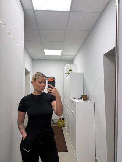 A woman is taking a selfie in a room with a dropped tile ceiling. The woman is wearing a black t-shirt and black sweatpants. Her hair is blonde and pulled back. She is holding an iPhone with an orange-accented case. The room has a neutral, office-like feel. The image does not contain location information for Sarapul, Russia.