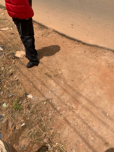 The image shows a person standing on a dirt path next to a paved road. The person is wearing a red puffer jacket and black pants with a chain attached to one of the belt loops. They are also wearing black sandals. The ground is covered in dry grass and some litter. The paved road appears to be the main road in Du, Nigeria. The sun is shining, casting shadows on the ground.