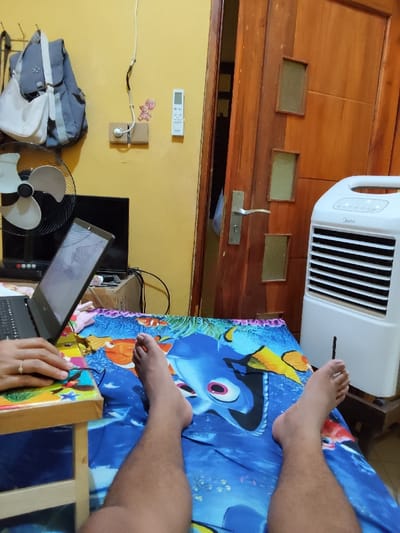 The image shows a person lying on a bed with a blue and white Finding Nemo themed sheet. The person's feet are visible and both are bare. The person is wearing a silver ring on their left hand. To the left of the bed is a wooden table with a black laptop resting on it. The screen of the laptop is open and visible. A black fan is behind the table. A white and grey electric fan is in the background on the right side of the image. The room has yellow walls. The image was taken at a location in Sidoarjo, Indonesia.
The coordinates given are: Latitude -7.448, Longitude: 112.714.
