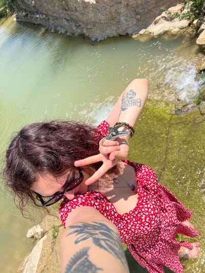 A woman is lying on the edge of a natural rock formation above a stream, possibly in or near Picassent, Spain, based on the information given. She is wearing a red, floral-patterned dress and black-rimmed glasses. The woman has dark, curly hair and is showing a peace sign with her left hand, which also has keys hanging from a ring. Tattoos are visible on both arms. Below her is a shallow stream with light-colored water flowing over rocks. The surrounding area appears to be rocky and natural.