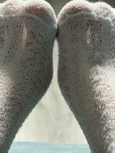 The image is a close-up, low-angle shot of two feet wearing white, sheer, patterned socks. The socks have small, diamond-shaped cutouts and are somewhat translucent, allowing the skin beneath to be visible. Sunlight is streaming in from the left, creating highlights and shadows on the socks and casting a light on the background. The feet are positioned close together, with the toes pointing slightly upwards and towards the viewer. The surface beneath the feet appears to be a light blue or turquoise color, possibly a table or floor. The background is out of focus, showing a lighter, neutral color. There is no indication of a specific location in Naberezhnye Chelny, Russia, or any context beyond the close-up view of the socks and feet.