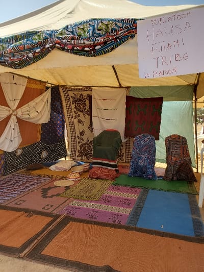 The image depicts the interior of a white canopy tent in Bar Gada, Nigeria, serving as an exhibition or display area. A white sign handwritten with "10 PLATOON HAUSA FULANI TRIBE 10 PLATOON" hangs from the tent's internal frame. The tent walls are draped with various patterned textiles, including white embroidered fabric, dark blue geometric prints, beige floral patterns, and dark red/green designs. A colorful patterned fabric valance adorns the front upper opening. The floor is covered by an arrangement of interlocking rectangular woven mats in orange, pink, blue, green, and brown hues. Positioned on the floor towards the rear are a light-colored, possibly embroidered cushion, and two circular woven caps. Three upright figures, resembling mannequins, are draped in distinct patterned fabrics: one in dark green with red and black horizontal stripes, another in blue patterned textile, and the third in brown geometric patterned fabric. No individuals are present in the scene.