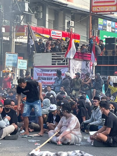 The image captures an outdoor public gathering or protest occurring on an asphalt road in Pati, Indonesia, under clear daylight conditions.

In the foreground, a bare-chested adult male, exhibiting a large build and covered in a pale substance (possibly powder or paint), sits directly on the ground wearing light-colored shorts and green flip-flops. He holds a long wooden stick across his lap. Standing nearby, also barefoot, is another adult male dressed in a black t-shirt and ripped blue denim shorts, looking downwards. Surrounding these individuals, numerous other people are seated or standing on the asphalt, attired in casual clothing, caps, and some wearing face masks.

In the midground, a red flatbed truck functions as an elevated platform, densely occupied by individuals. Multiple flags are displayed from the truck, including a black flag with a white skull and crossbones (Jolly Roger) and several red-and-white Indonesian national flags. Large sound system speakers are positioned on the right side of the truck bed. A prominent white banner below the truck reads "TUNDUR DITINDAS ATAU BANGKIT ME..." and "SEBAB DIAM ADALAH PENGKHIAN..." (translating from Indonesian to "FALL UNDER OPPRESSION OR RISE UP..." and "BECAUSE SILENCE IS TREASON..."). A partial banner displaying "{KELOMPOK. P..." is also visible.

The background features a multi-story building with an "Alfamart" sign and various electronic brand advertisements (Acer, Asus, Epson, MSI). Multiple air conditioning units are mounted on the building's exterior. Overhead power lines and road signs are present on the left side of the frame. Smoke or haze is noticeable in the upper left background. Scattered litter, including plastic bottles and footwear, is visible on the asphalt.