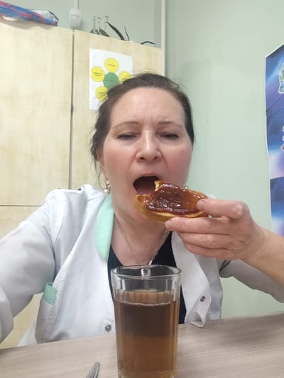 A woman is indoors, seated at a table, holding a piece of bread spread with a brown topping, seemingly jam, near her open mouth, as if about to eat it. In front of her on the table is a glass filled with a dark-colored liquid. She is wearing a white coat with a light green trim, suggesting a professional setting, possibly a doctor's office or clinic. Her hair is pulled back, and she is wearing earrings. The background features a light green wall, some light wood cabinets, and a poster with images related to space. The location is Sarapul, Russia.