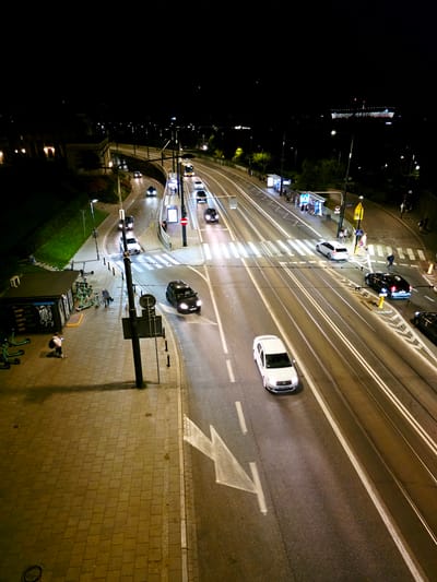 This is a high-angle, night-time shot of a street in Warsaw, Poland. The scene depicts a busy intersection with traffic, including cars and what appear to be trams or streetcars, indicated by the tracks. The time of day is clearly night, as evidenced by the darkness and the artificial lighting from streetlamps, car headlights, and illuminated signs. The weather appears to be clear.

Several cars are visible on the road, some with their headlights on, creating light trails due to the long exposure. A white car is in the foreground, moving to the right. Further up the road, a black car is also in motion. There are also pedestrian crossings marked on the road, with white stripes.

On the left side of the frame, there's a darker area with trees and more streetlights. On the right, a pedestrian walkway made of brick paving stones is visible. A few people are present in the scene. One person is standing on the walkway, and others are near a bus or tram stop, which has illuminated shelters. A few people are also walking across the street on a pedestrian crossing.

There are some visible text elements, including signage at the bus stop and some writing on a dark structure in the lower left, which appears to be a piece of street art or advertisement. The overall impression is a dynamic urban environment at night, with the movement of vehicles and the presence of pedestrians.