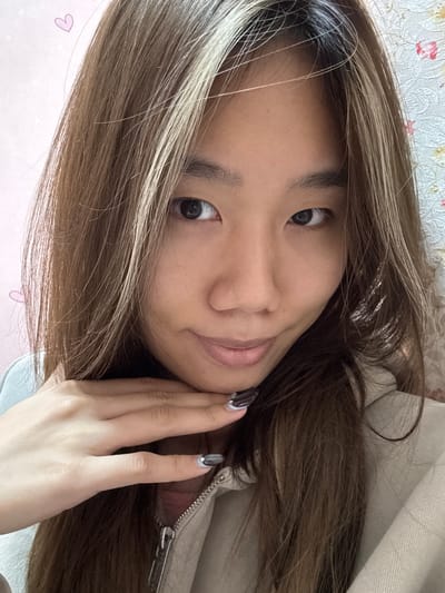 A young Asian woman poses for a selfie. She has long, brown hair with blonde highlights. She wears a cream-colored hoodie. Her left hand is touching her chin. She has painted nails with a silver and white design. The background shows a bed with a light pink and white floral pattern. There are pink heart patterns on the bed. The photo may have been taken in Shapingba District, China.