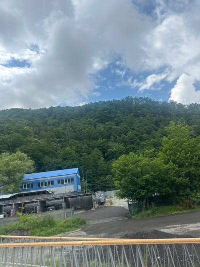 A wide shot shows a blue-roofed industrial building situated at the base of a densely forested, mountainous slope. The sky above is filled with dramatic, white cumulus clouds against a backdrop of blue. In the foreground, a car wash facility with multiple bays is visible, with cars and workers present. A grey metal railing with a wooden top runs across the bottom of the frame, obscuring part of the car wash and the ground. The scene appears to be set in Estosadok, Russia, a resort town known for its natural beauty and proximity to mountains.