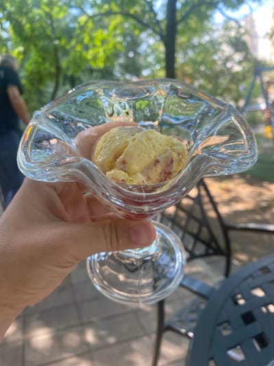 The image shows a hand holding a clear glass dish filled with ice cream. The ice cream is light yellow with visible red chunks, possibly fruit or candy. The glass dish has a wavy, decorative rim. In the background, out of focus, are green trees, suggesting an outdoor setting with dappled sunlight. A portion of a grey, decorative metal table or chair is also visible. The overall impression is one of a casual, sunny day, likely outdoors, enjoying a dessert. The scene evokes a sense of relaxation and enjoyment. No text is visible in the image.