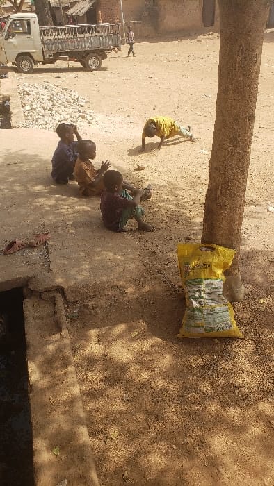 The image depicts an outdoor scene in Kaduna South, Nigeria, under direct sunlight, featuring an unpaved dirt ground. In the immediate foreground on the right, a thick brown tree trunk stands, with a crumpled yellow sack, bearing green and white markings, resting against its base. Adjacent to the tree, a dark-skinned child in a yellow shirt and blue pants is positioned prone on the ground, supported by hands and knees, facing left. Three other dark-skinned children are seated on the dirt ground to the left of the prone child: one in a dark blue garment with hands near their head, one in a light brown top with hands clasped, and one in a dark red top and green pants holding a dark rectangular object. All three seated children face towards the right. To the lower left, a pair of red plastic sandals lies on a concrete slab that partially covers an open dark drain. In the background, a light-colored pickup truck with a utility bed is parked on the left, near a pile of light-colored rubble. Further back, structures with light brown walls are visible. An individual in a plaid shirt and dark pants walks from right to left across the distant background.