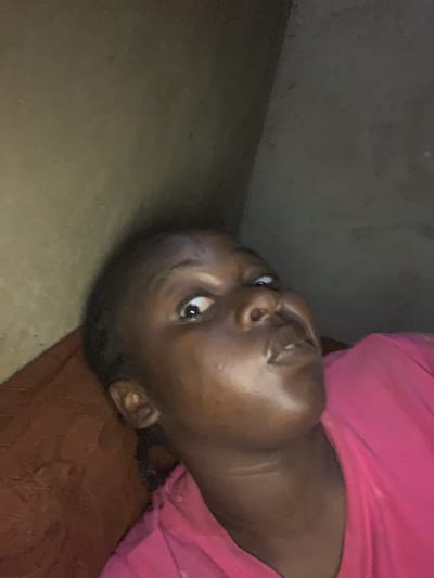 A close-up photograph shows a dark-skinned person lying on a surface, possibly a bed or couch, with their head tilted towards the left of the frame. They are wearing a pink shirt. The background includes a beige wall and brown surface. The location is not discernible as Agban, Nigeria.