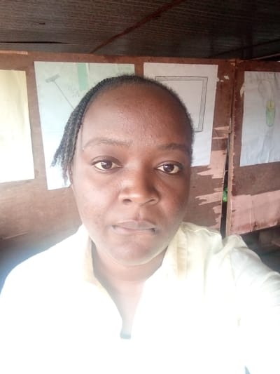 This is a medium close-up selfie of a Black woman in Ukwu-Abwa, Nigeria. She has dark skin and her hair is braided and pulled back. She is wearing a light yellow collared shirt. The background appears to be a room with wooden walls and what looks like a ceiling made of corrugated metal. There are several pieces of paper taped to the wall, which seem to be children's drawings. One drawing clearly depicts a rake. The lighting suggests it is daytime, possibly indoors with natural light contributing to the overall brightness. The woman's expression is neutral.